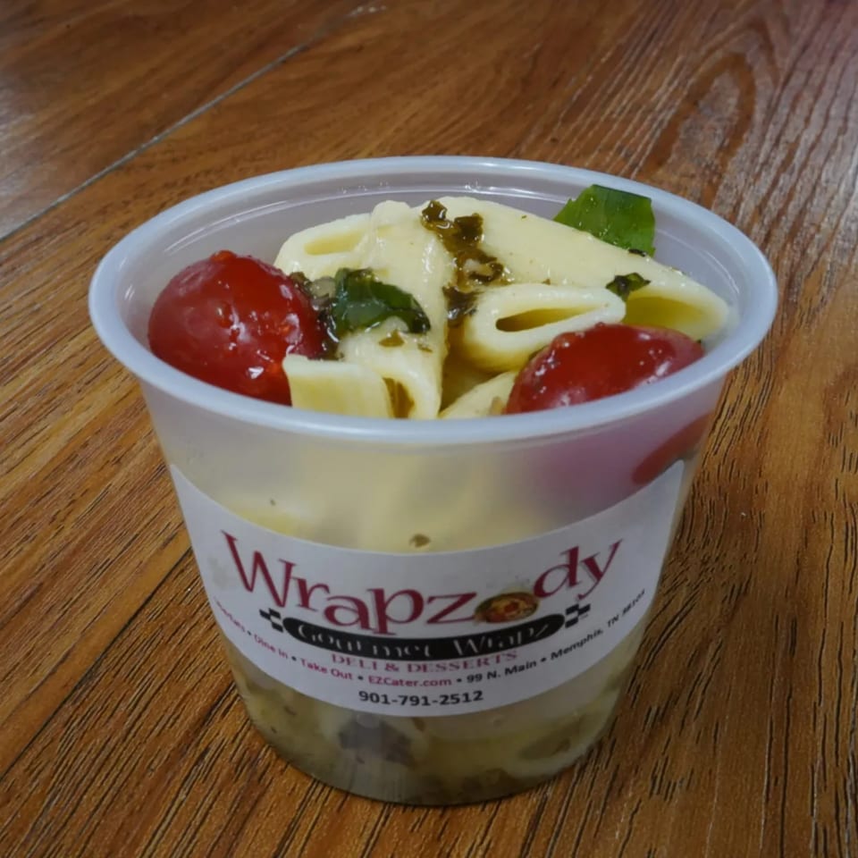 Wrapzody's pasta salad with cherry tomatoes and herbs in a clear plastic cup on a wooden table.