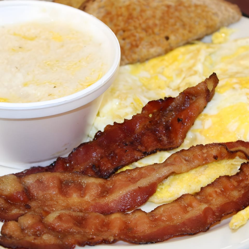 A breakfast plate with scrambled eggs, crispy bacon strips, a side of grits, and toast.