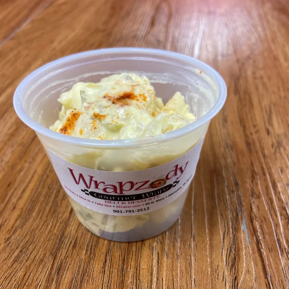 A clear plastic cup of potato salad with paprika on top, from Wrapzody Gourmet Wrapz Deli.