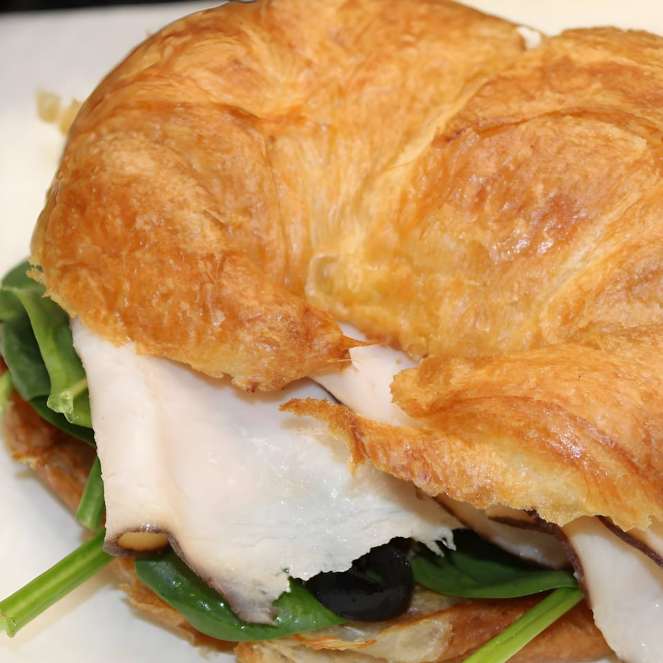 A golden-brown croissant sandwich filled with white meat, green spinach, and black olives.